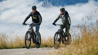 Kaksi pyöräilijää ajaa e-mountainbikeillaan pyörätiellä. Taustalla on vuoria.